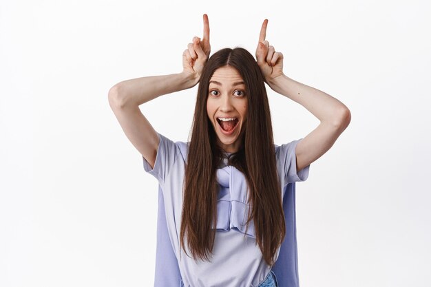 Cerca de divertida chica positiva, haciendo toros cuernos de diablo con los dedos en la cabeza, sonriendo emocionado, de pie contra la pared blanca