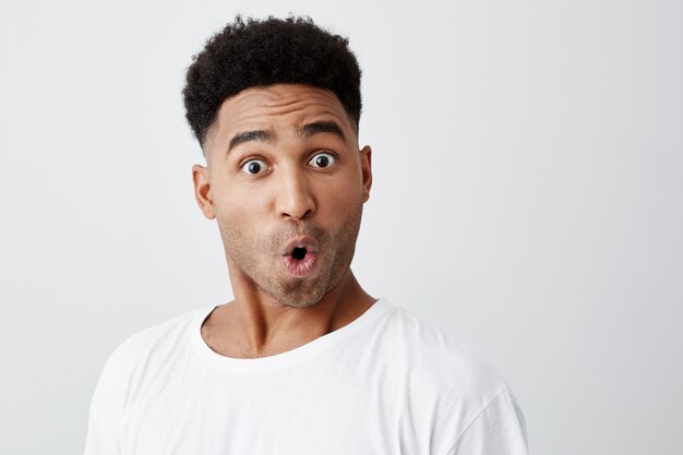 Cerca de divertido guapo joven de piel oscura con corte de pelo afro en elegante camiseta blanca mirando a la cámara con las cejas levantadas y la expresión de la cara sorprendida.