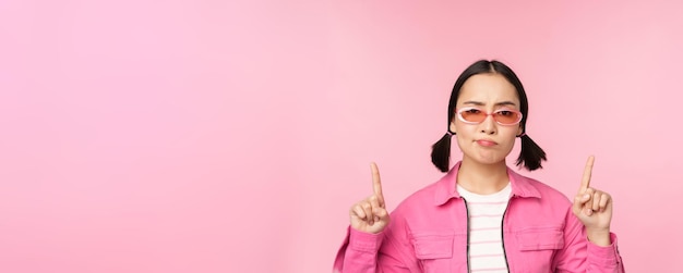 Cerca de una elegante chica coreana que parece perpleja señalando con el dedo hacia arriba mostrando un anuncio de pie sobre un fondo rosa