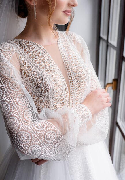 Cerca del elegante vestido de macramé blanco usado por la novia de pie junto a la ventana