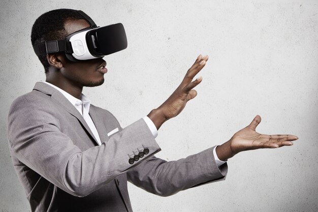 Cerca de un empleado africano con traje formal y gafas, experimentando la realidad virtual, estirando los brazos como si sostuviera algo con las manos.