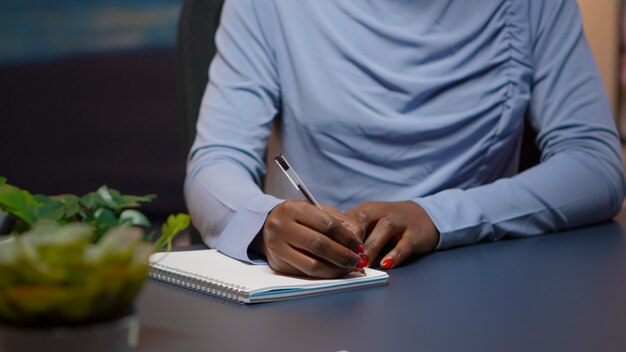 Cerca de la empresaria africana escribiendo la lista de tareas para el proyecto empresarial en el cuaderno sentado en el escritorio en la sala de estar trabajando horas extraordinarias. Freelancer negro respetando la fecha límite estudiando a altas horas de la noche