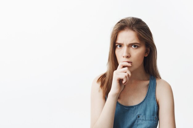 Cerca de la encantadora y encantadora joven europea con cabello largo y oscuro en ropa casual azul de la mano cerca de los labios, con expresión pensativa tratando de recordar el nombre de la persona