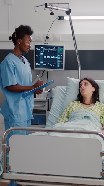 Cerca de la enfermera afroamericana monitoreando a la mujer enferma mientras descansa en la cama en la sala del hospital