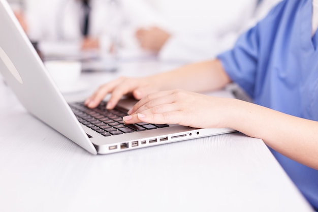 Cerca de la enfermera médica escribiendo en la computadora portátil durante la reunión de salud con compañeros de trabajo en la sala de juntas del hospital. Terapeuta experto de la clínica hablando con colegas sobre la enfermedad, profesional de la medicina