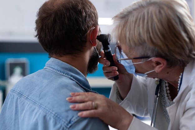 Cerca del especialista con otoscopio para hacer un examen de oído con el paciente. Mujer otóloga que comprueba la infección con un instrumento de otorrinolaringología en una visita médica durante la pandemia de coronavirus.
