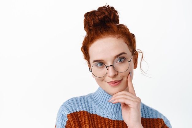 Cerca de estudiante pelirroja con moño, con gafas, tocando la mejilla y mirando pensativo, tiene una idea interesante, de pie sobre fondo blanco.