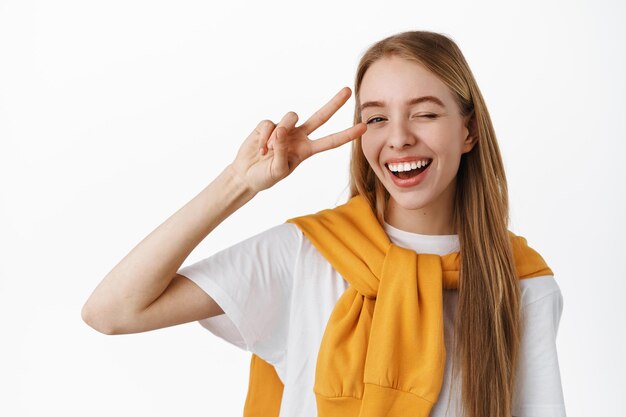 Cerca de feliz mujer caucásica con cabello lacio rubio, guiñando un ojo positivo y sonriendo, mostrando el gesto de paz v-sign cerca del ojo, de pie contra el fondo blanco