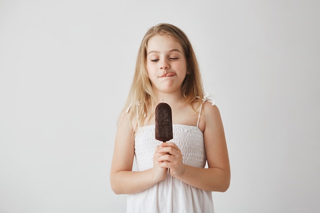 Cerca de graciosa niña con cabello largo claro en vestido blanco mostrando la lengua, con helado en las manos, no puedo esperar para comerlo.