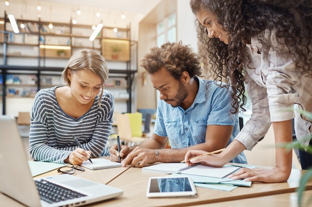 Cerca de un grupo de jóvenes iniciadores sentados en la biblioteca haciendo investigaciones sobre proyectos futuros, mirando a través de gráficos en la computadora portátil, escribiendo nuevas ideas.