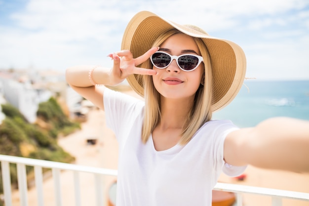 Cerca de la hermosa joven con sombrero de verano tomando un selfie y mostrando un gesto de paz en la playa