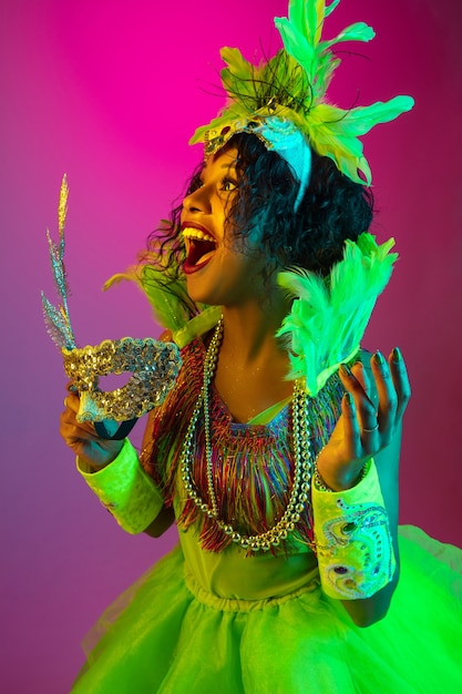 De cerca. Hermosa mujer joven en carnaval, elegante disfraz de mascarada con plumas en pared degradada en luz de neón. Concepto de celebración navideña, tiempo festivo, baile, fiesta, diversión.