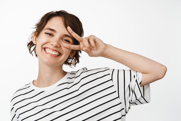 Cerca de la hermosa niña feliz, sonriendo dientes blancos, mostrando el signo v de paz cerca del ojo, expresión de la cara alegre positve, de pie en blanco