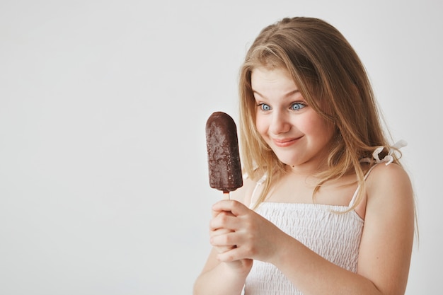 Cerca de hermosa niña pequeña de pelo claro con ojos azules en vestido blanco con helado en las manos, mirándolo con expresión de cara feliz.