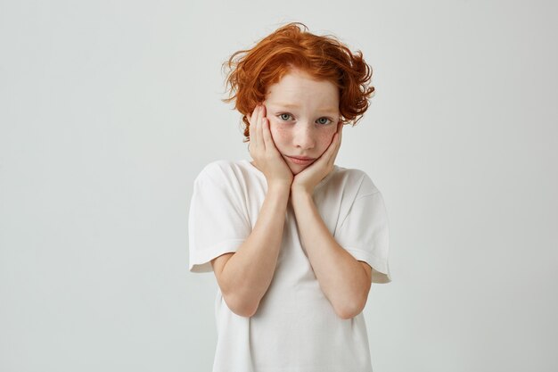 Cerca de hermoso niño pequeño con pelo de jengibre y pecas cogidos de la mano en las mejillas, mirando, estar cansado