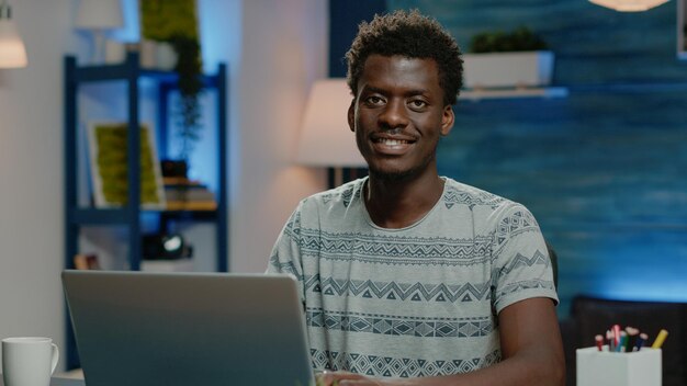 Cerca del hombre afroamericano trabajando en equipo portátil. Empleado negro haciendo trabajo remoto en internet mientras mira a cámara y sonríe. Persona de negocios de etnia africana con dispositivo