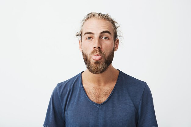 Cerca de hombre guapo divertido con buen peinado y barba en camiseta azul haciendo cara tonta