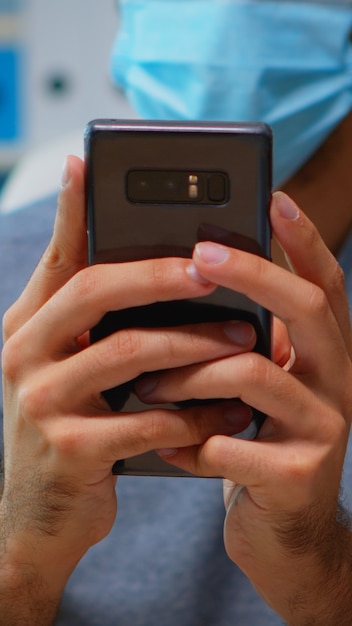 Foto gratuita cerca del hombre con mascarilla escribiendo mensajes de texto en el teléfono inteligente sentado en el lugar de trabajo durante el coronavirus. freelancer que trabaja en la nueva oficina normal charlando hablando por escrito utilizando tecnología de internet