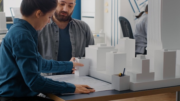 Cerca de un hombre y una mujer que diseñan el plan de planos, analizan el modelo de construcción. Equipo de personas que trabajan en el diseño de la construcción con boceto industrial y maqueta para proyecto urbano.