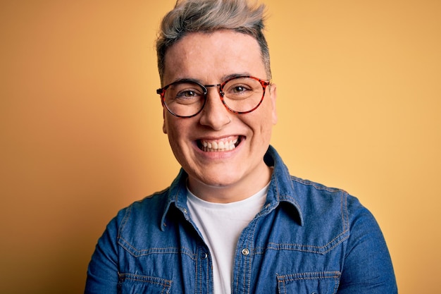 Cerca de un joven apuesto y moderno con gafas y chaqueta vaquera sobre fondo amarillo con una cara feliz de pie y sonriendo con una sonrisa segura mostrando los dientes