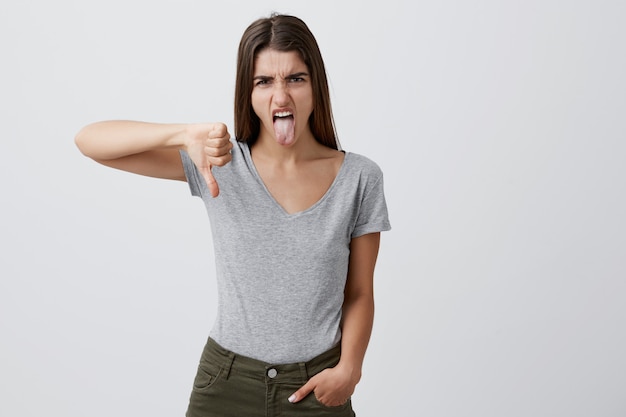 Cerca de la joven y atractiva chica caucásica negativa atractiva con cabello largo y oscuro en camiseta gris y jeans mostrando el pulgar hacia abajo y la lengua con expresión infeliz y mala,