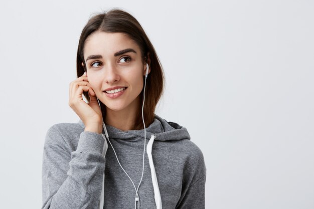 Cerca de la joven y atractiva mujer caucásica alegre con cabello oscuro en sudadera con capucha gris informal sonriendo alegremente, sosteniendo los auriculares con la mano, mirando a un lado con expresión de la cara soñadora y alegre.