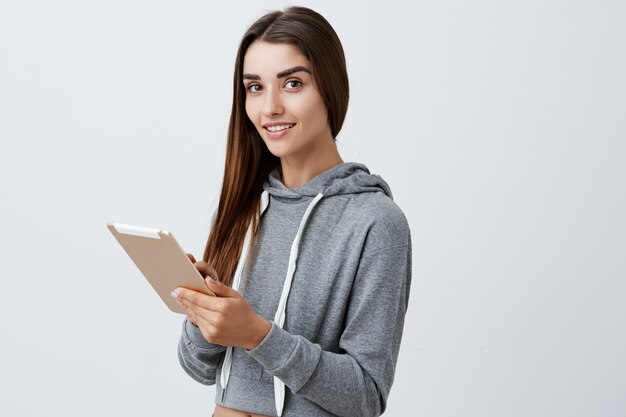 Cerca de la joven y bella muchacha alegre caucásica con cabello largo y oscuro en traje deportivo casual sonriendo brillantemente chateando con amigos en tableta digital