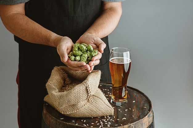 Foto gratuita cerca del joven cervecero confiado con cerveza artesanal en vidrio en barril de madera en la pared gris.