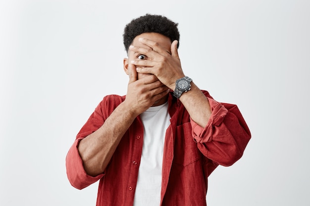 Cerca del joven hombre de piel oscura con peinado afro en camiseta blanca bajo camisa roja ropa cara con las manos, mirando a través de los dedos con expresiones de cara asustada.