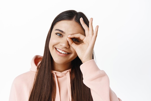 Cerca de una joven mujer feliz haciendo un buen signo en el ojo, diga sí, elogie algo bueno de pie contra el fondo blanco