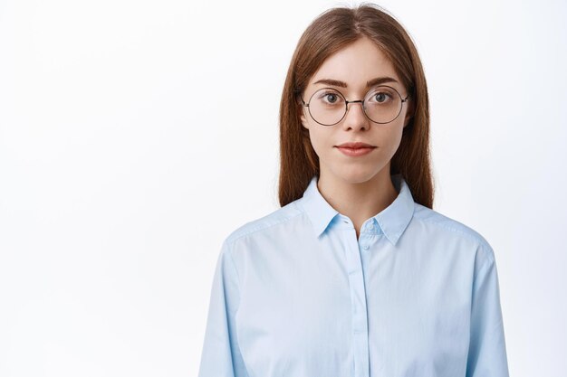 Cerca de la joven mujer de oficina en camisa azul de negocios y gafas, con aspecto de profesional con rostro decidido, de pie sobre fondo blanco.