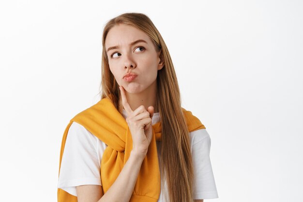 Cerca de una joven pensativa, haciendo una cara pensativa, tocando la barbilla y los labios fruncidos, mirando la promoción del producto de la esquina superior derecha mientras piensa, toma una decisión, de pie sobre una pared blanca