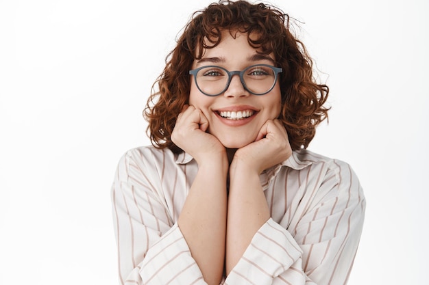 Foto gratuita cerca de una joven sonriendo complacida, mirando algo hermoso, mirando con admiración, usando anteojos, admirando o soñando despierto, de pie sobre fondo blanco