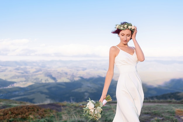 Cerca de joven con un vestido blanco con una corona en la cabeza y un ramo de flores al aire libre