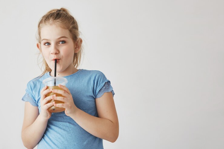 Cerca de linda chica con cabello claro en peinado de cola con vaso de leche en las manos, bebiendo a través de paja y mirando a un lado con cara divertida.