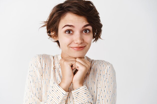 Cerca de linda y tierna chica morena en blusa con aspecto romántico, sonriendo y mirando coqueta, de pie contra la pared blanca