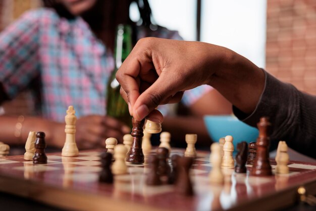 Cerca de la mano del hombre moviendo la pieza de ajedrez de la reina mientras jugaba en casa con amigos. Persona inteligente que disfruta del juego de mesa de estrategia mientras se sienta a la mesa en la sala de estar con un grupo multiétnico de personas.