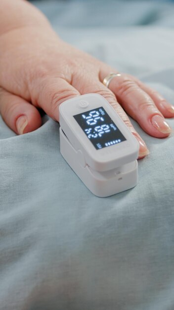 Cerca de la mano del paciente anciano con oxímetro en la cama. Persona mayor con herramienta médica en el dedo para medir la saturación de oxígeno y la presión del pulso. Pensionista con enfermedad