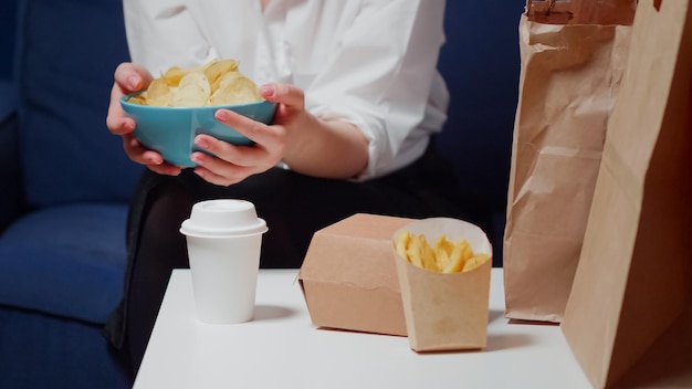 Cerca de las manos caucásicas sosteniendo el cuenco de patatas fritas sentado en el sofá. Mujer joven con comida para llevar en la mesa y bocadillos poco saludables preparándose para comer en la sala de estar en casa después del trabajo