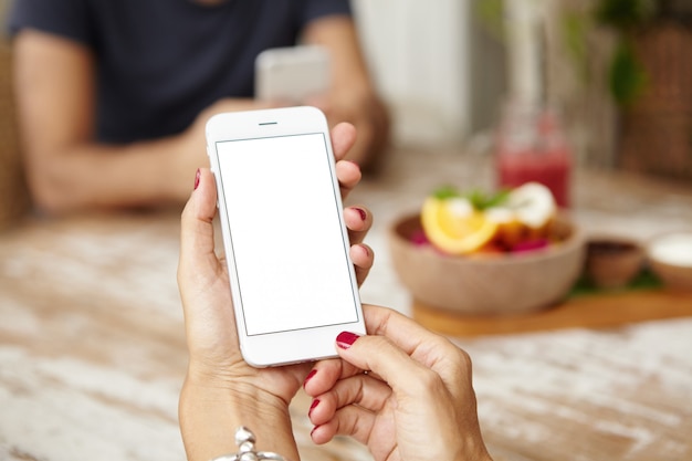 Cerca de manos femeninas con uñas rojas sosteniendo teléfono inteligente con pantalla de espacio de copia en blanco para su texto o contenido publicitario. Mujer caucásica navegando por internet en el teléfono celular durante el almuerzo.