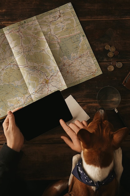 Cerca de las manos del hombre sosteniendo la tableta y deslizándose con el dedo, planeando una ruta de aventura en una mesa de madera envejecida con un mapa de carreteras mientras el curioso perro basenji lo mira con las patas en la mesa