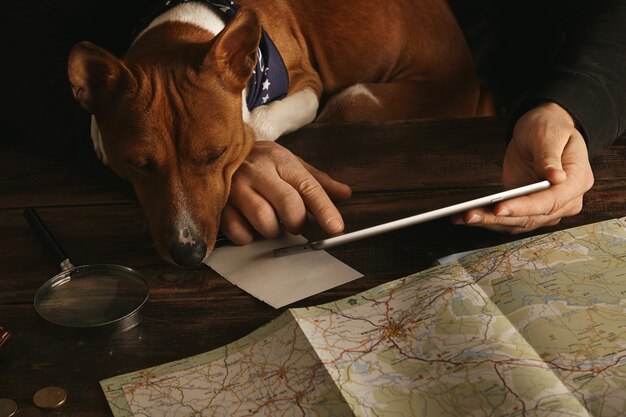 Cerca de las manos del hombre sosteniendo la tableta y deslizándose con el dedo, planeando la ruta de la aventura en la mesa de madera envejecida mientras el curioso perro basenji lo mira con las patas en la mesa