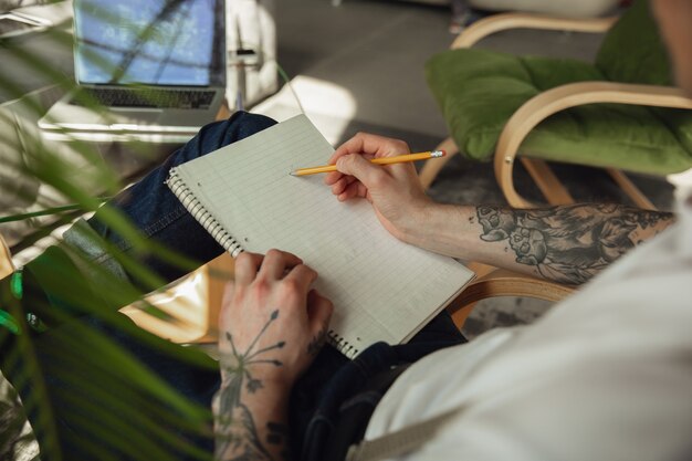 Cerca de manos masculinas escribiendo en un papel vacío sobre la mesa en casa.