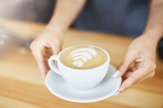 Cerca de manos de mujer sirviendo un capuchino en una taza con arte latte. Concepto de barista.