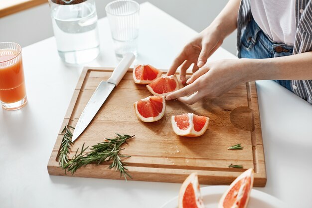 Cerca de las manos de la niña en rodajas cuchillo de pomelo y romero en el escritorio de madera. Copia espacio