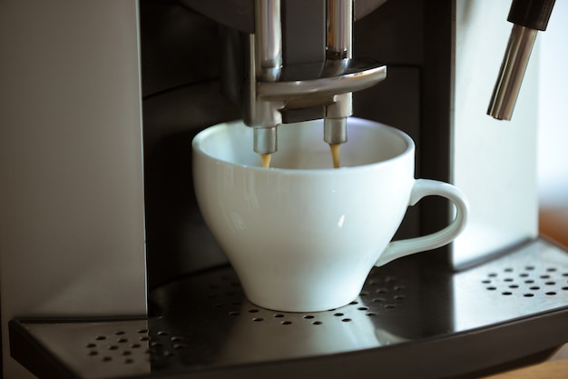Cerca de la máquina de café vertiendo capuchino, espresso, americano en taza blanca en casa o café. Bebida caliente sabrosa y aromática. Comida, nutrición, la bebida más popular para el desayuno y el descanso en el horario laboral.