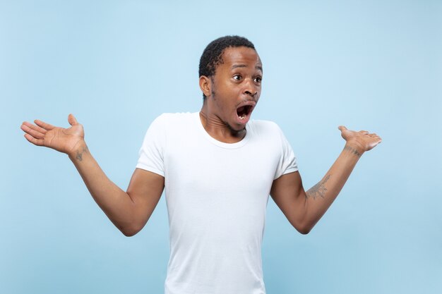 Cerca de medio cuerpo retrato de joven afroamericano con camisa blanca en el espacio azul. Emociones humanas, expresión facial, publicidad, concepto de ventas. Invitando, parece sorprendido y asombrado