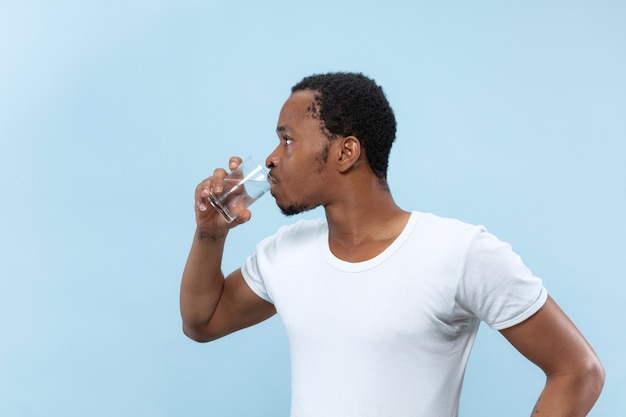 Cerca de medio cuerpo retrato de joven afroamericano con camisa blanca en la pared azul. Las emociones humanas, la expresión facial, el concepto publicitario. Sosteniendo un vaso y agua potable.