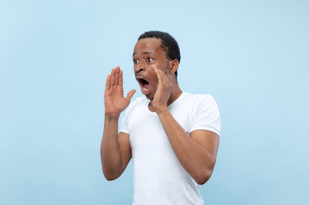 Cerca de medio cuerpo retrato de joven afroamericano con camisa blanca en la pared azul. Emociones humanas, expresión facial, publicidad, ventas, concepto. Gritando, llamando a alguien, anuncia.