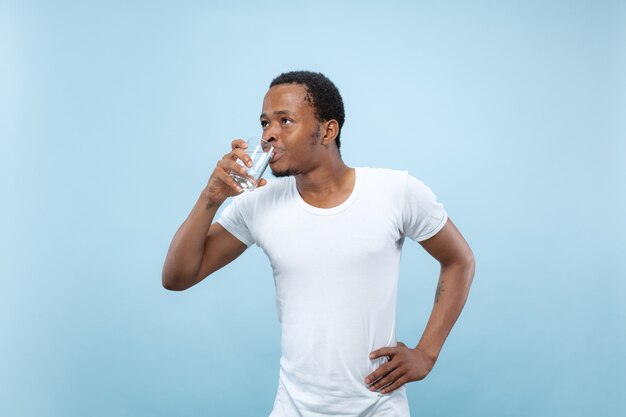 Cerca de medio cuerpo retrato de joven afroamericano con camisa blanca sobre fondo azul. Las emociones humanas, la expresión facial, el concepto publicitario. Sosteniendo un vaso y agua potable.